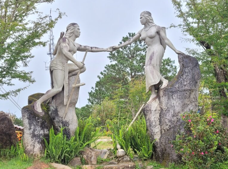 Tượng đôi trai gái Lang và Biang đứng bên nhau trên đỉnh núi, phía sau là rừng thông xanh.