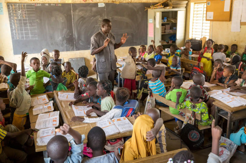 Giáo viên và học sinh trong lớp học nhỏ ở vùng nghèo, bàn ghế bằng gỗ thô sơ nhưng tràn đầy niềm vui.